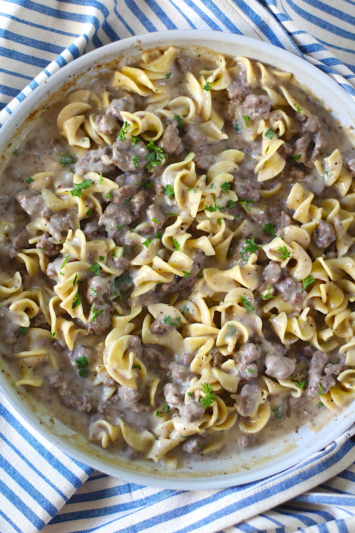 Creamy Beef and Noodles with ground beef and parsley garnish in a skillet on table on blue and white striped towel.