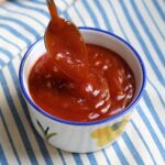 Spoon scooping Honey Sriracha Sauce in a small bowl on a blue and white striped tablecloth.