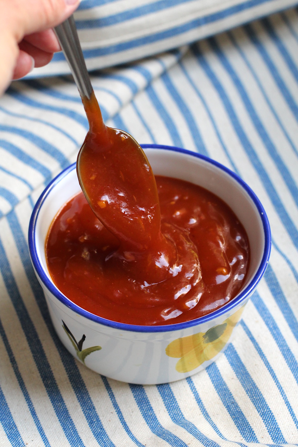 Spoon scooping Honey Sriracha Sauce in a small bowl on a blue and white striped tablecloth.