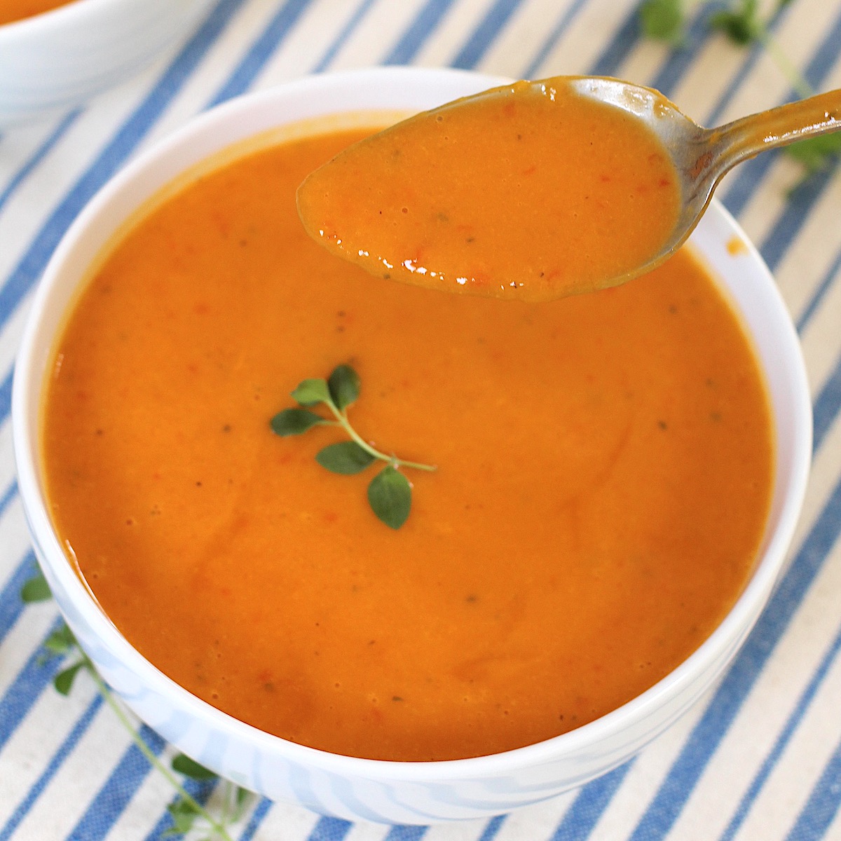 Spoon holding spoonful of Sweet Potato and Red Pepper Soup over the bowl garnished with fresh oregano leaves. Blue and white striped tablecloth in background.