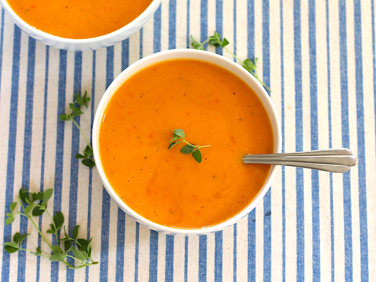 Sweet Potato and Red Pepper Soup in two bowls with fresh oregano leaves around the table and garnishing the soup in one bowl. Blue and white striped tablecloth in background.