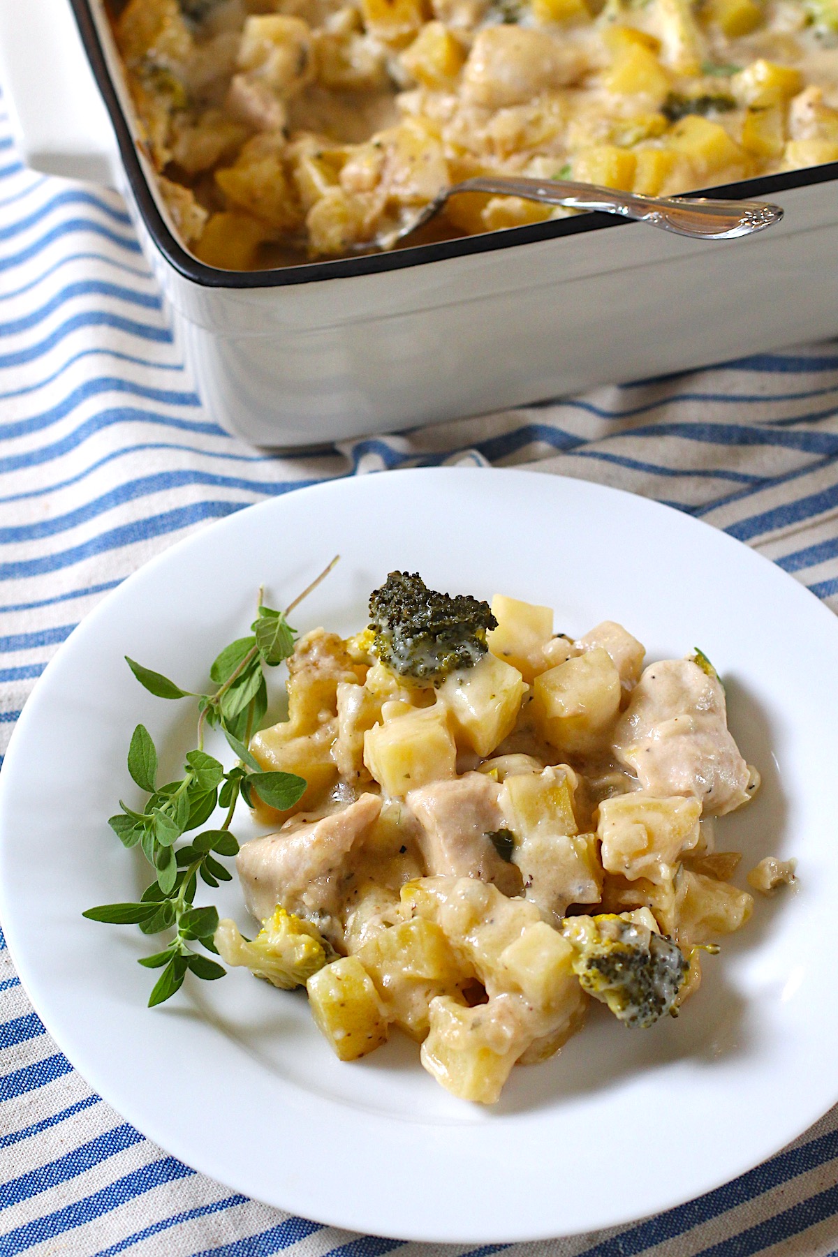 Chicken Broccoli Potato Casserole on a plate with a sprig of fresh oregano and the casserole dish in the background, all on a blue and white striped tablecloth.