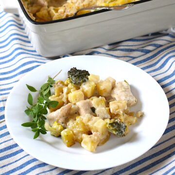 Chicken Broccoli Potato Casserole on a plate with a sprig of fresh oregano and the casserole dish in the background, all on a blue and white striped tablecloth.