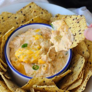 Slow cooker Chicken Buffalo Dip in a serving bowl with sliced scallions on top, tortilla chips all around the outside in another bowl, and a chip scooping dip from the bowl.