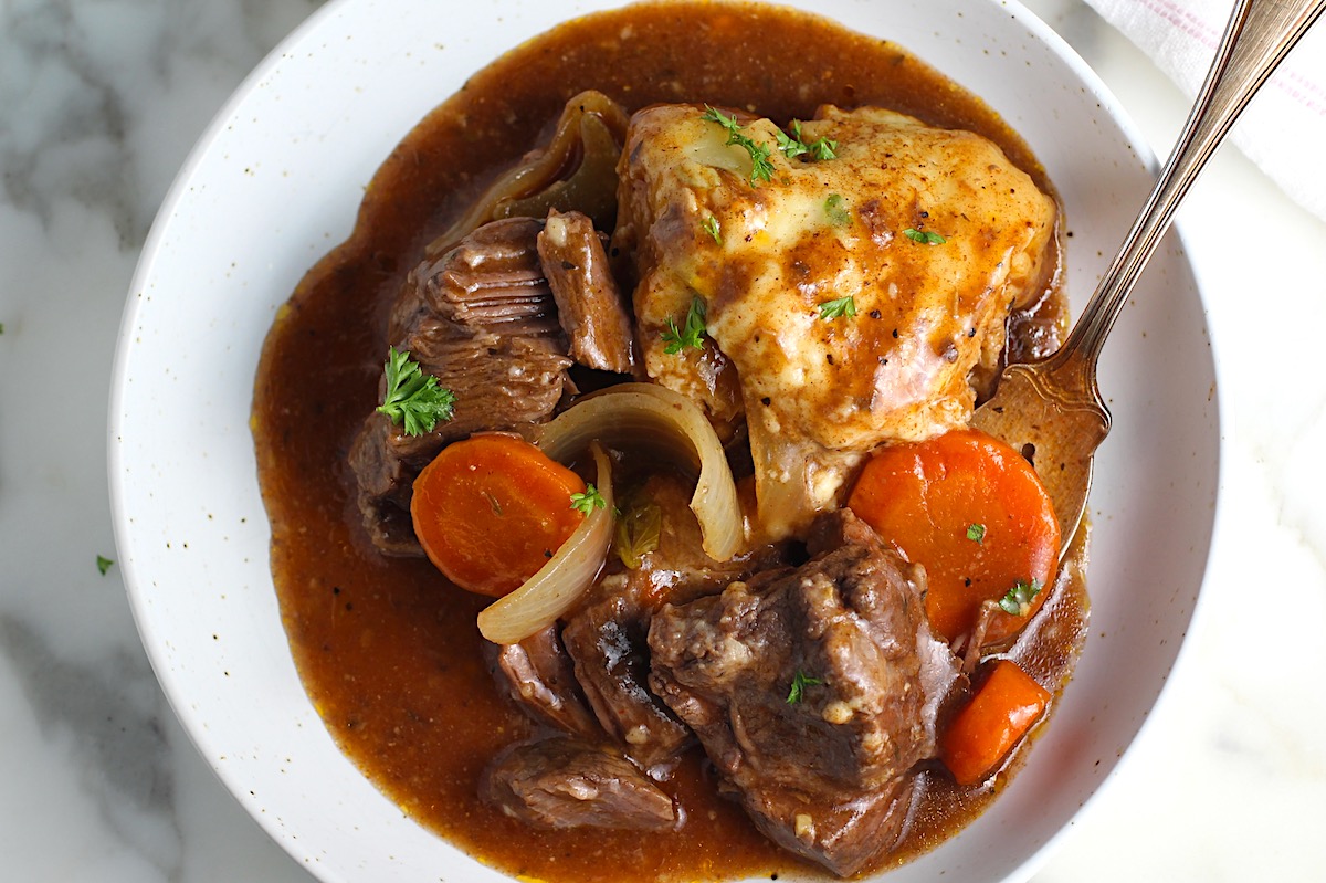 Crockpot Beef Stew with Dumpling in a bowl with carrots and onions and a fork on marble counter.
Parsley sprinkled on top.