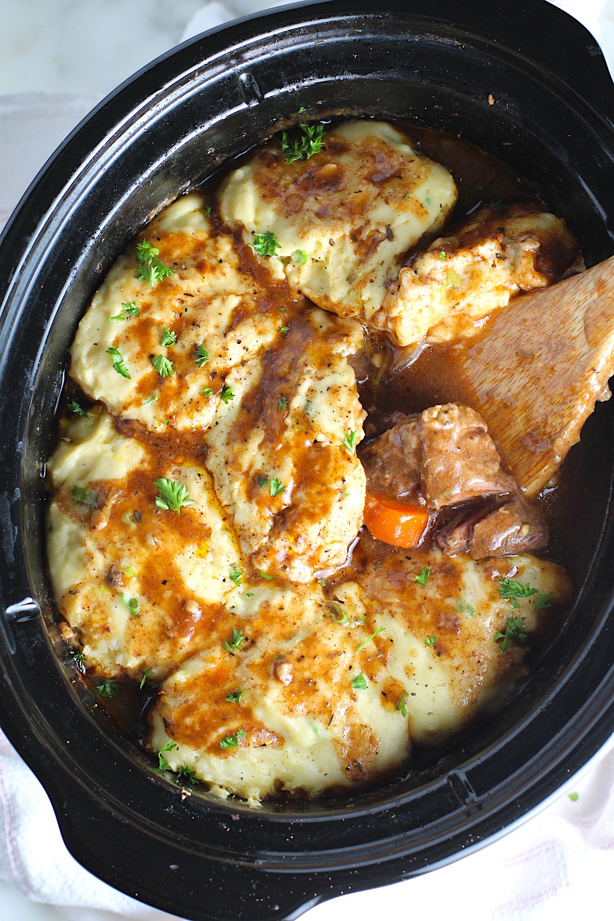 Crockpot Beef Stew with Dumplings on top in the crockpot with a wood spoon scooping tender beef cubes and carrots. Parsley sprinkled on top.