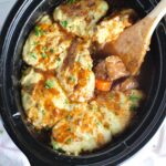 Crockpot Beef Stew with Dumplings on top in the crockpot with a wood spoon scooping tender beef cubes and carrots. Parsley sprinkled on top.