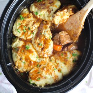 Crockpot Beef Stew with Dumplings on top in the crockpot with a wood spoon scooping tender beef cubes and carrots. Parsley sprinkled on top.