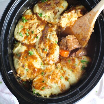 Crockpot Beef Stew with Dumplings on top in the crockpot with a wood spoon scooping tender beef cubes and carrots. Parsley sprinkled on top.