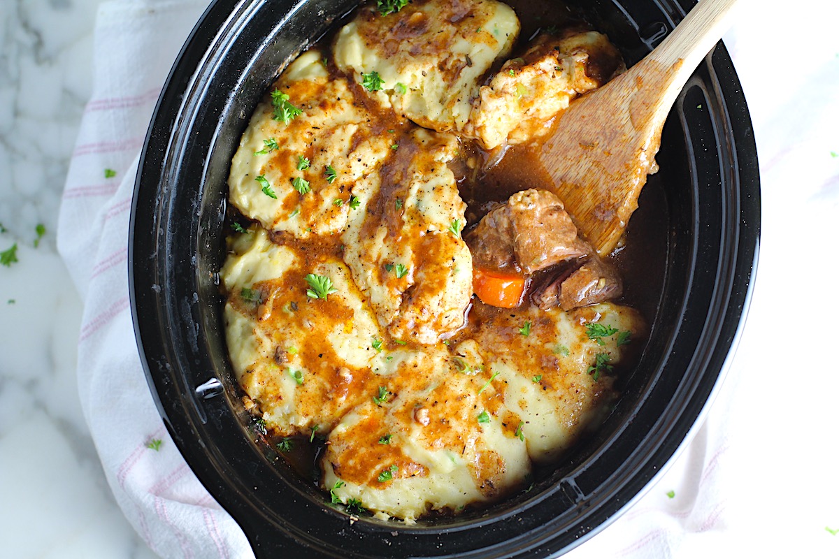 Crockpot Beef Stew with Dumplings on top in the crockpot with a wood spoon scooping tender beef cubes and carrots. Parsley sprinkled on top.