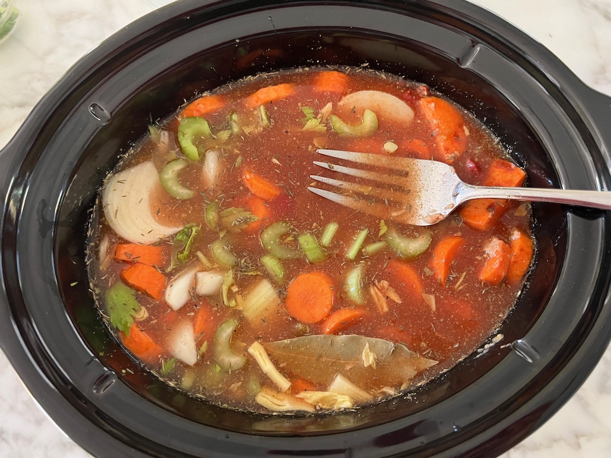 Fork mixing and making sure all ingredients are submerged in liguid after broth, veggies, and seasonings are added to the beef stew meat in the slow cooker.
