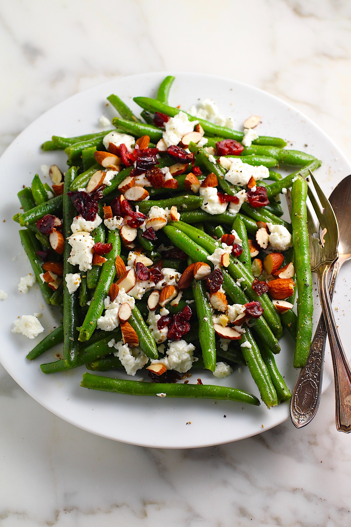 Whole Frozen Green Beans that have been sauteed in butter and garlic, then topped with goat cheese, almonds, and dried cranberries all on a platter with serving fork and spoon on the side.