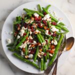 Whole Frozen Green Beans that have been sauteed in butter and garlic, then topped with goat cheese, almonds, and dried cranberries all on a platter with serving fork and spoon on the side.
