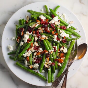 Whole Frozen Green Beans that have been sauteed in butter and garlic, then topped with goat cheese, almonds, and dried cranberries all on a platter with serving fork and spoon on the side.