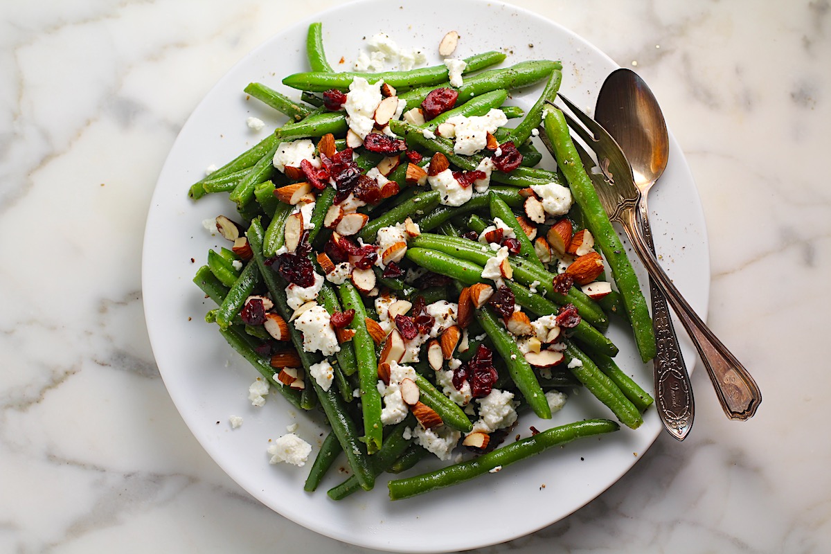 Whole Frozen Green Beans that have been sauteed in butter and garlic, then topped with goat cheese, almonds, and dried cranberries all on a platter with serving fork and spoon on the side.