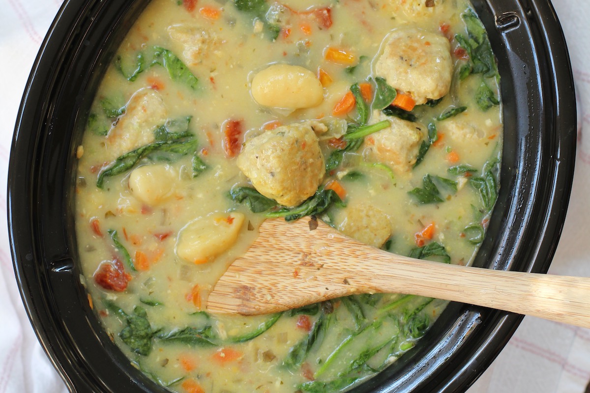 Slow Cooker Tuscan Chicken Meatballs with Gnocchi in creamy sauce with sun dried tomatoes and spinach in the slow cooker pot with wood spoon.