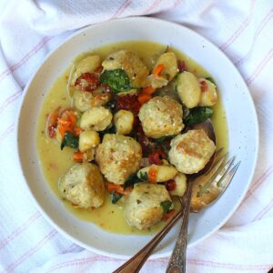Slow Cooker Tuscan Chicken Meatballs with Gnocchi in creamy sauce with sun dried tomatoes and spinach in a rimmed plate on counter with for and spoon on plate and the slow cooker pot background.