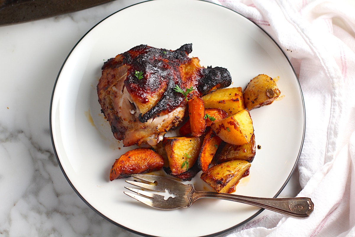 Tray baked Chicken Thighs, Potatoes, and carrots on a plate with fork perfectly browned glistening.
