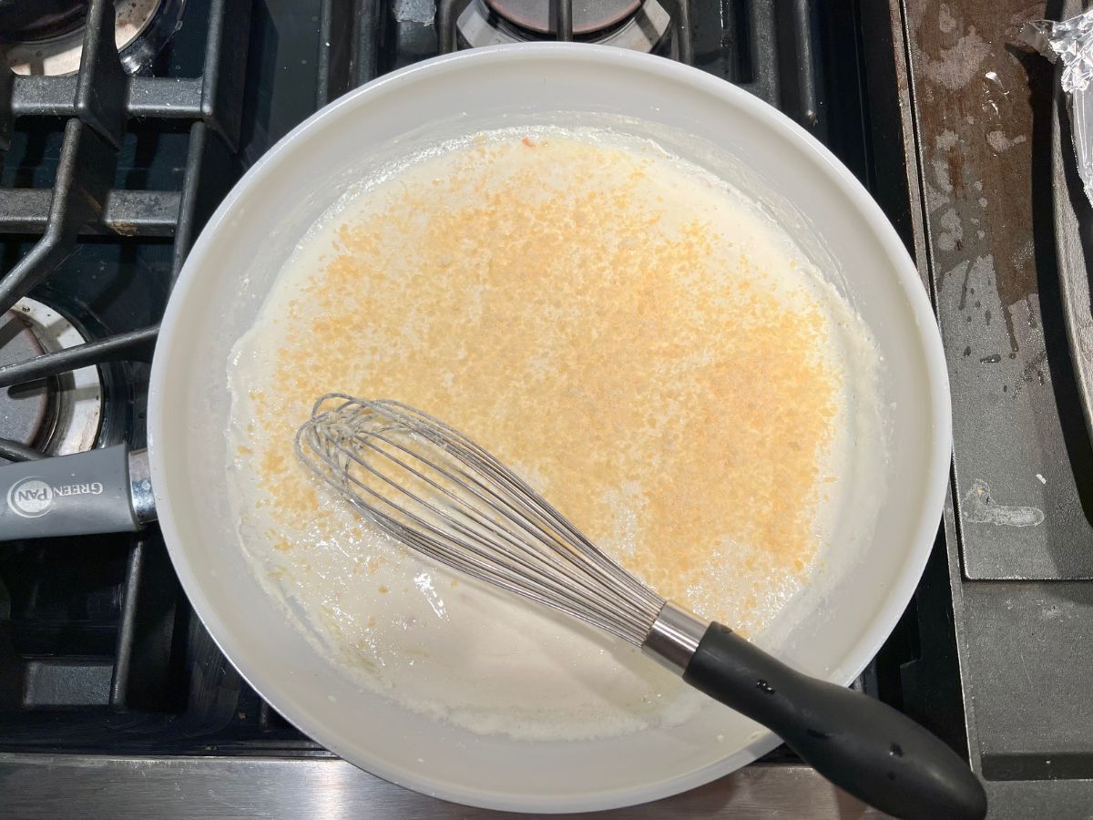 Whisk resting in a white sauce with parmesan cheese added to the skillet.