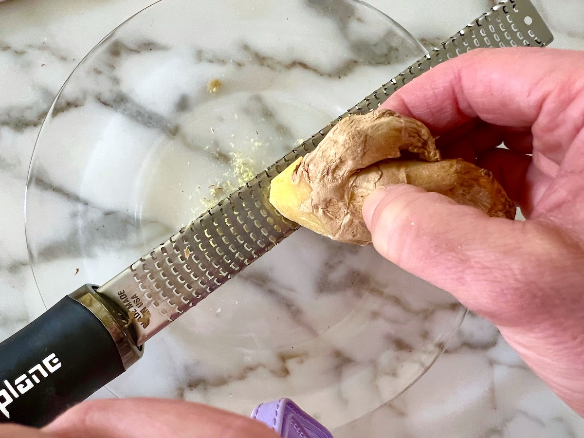 Hand holding ginger root over a microplane for Black Pepper Beef Stir Fry with onions.