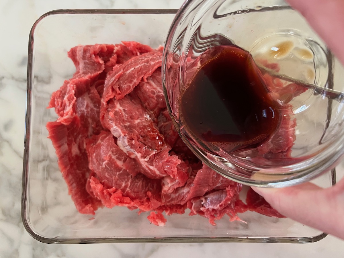 Marinade being poured over sliced raw flank steak in clear glass dish for Black Pepper Beef Stir Fry with onions.