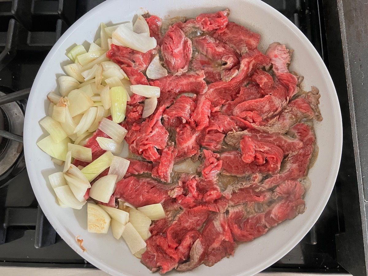 Raw flank steak slices, onions, and sauce cooking in pan for Black Pepper Beef Stir Fry with onions.