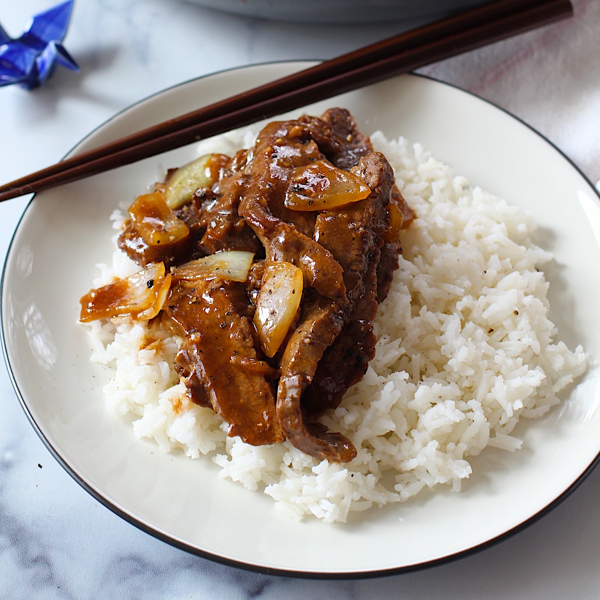 Black Pepper Beef Stir Fry with onions over white rice on a plate with chopsticks.