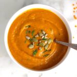 Creamy Carrot and Lentil Soup blended smooth in a bowl on counter with a spoon and garnished with pumpkin seeds, sunflower seeds, and parsley.