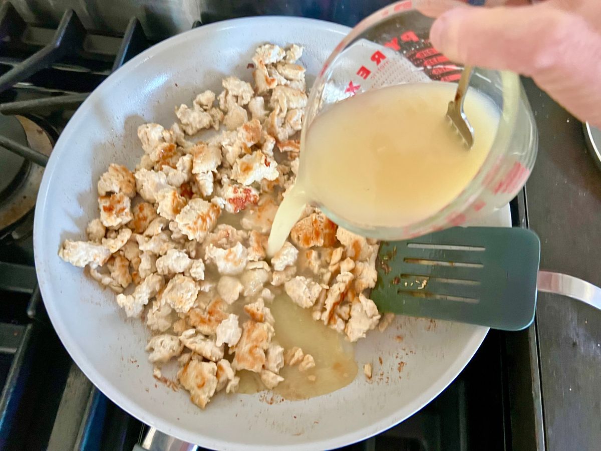 Chicken broth being poured into the skillet with ground chicken.