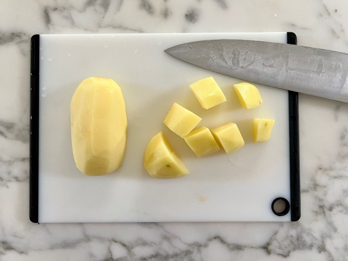Chef's knife resting on a cutting board next to a peeled potato that's partially diced.