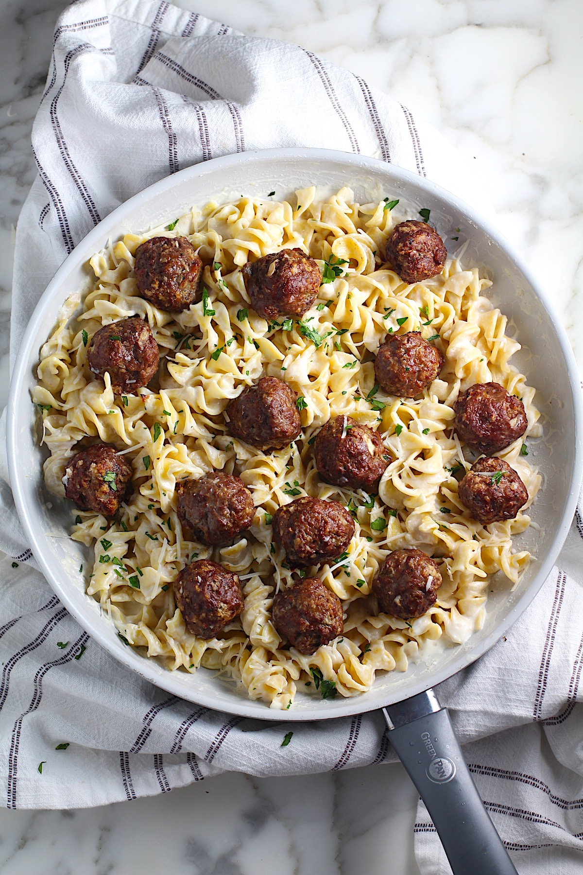 Alfredo and meatballs with meatballs nestled into creamy sauce and noodles in a frying pan on towel on counter.