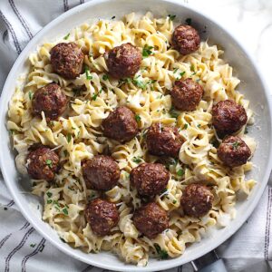 Alfredo and meatballs with meatballs nestled into creamy sauce and noodles in a frying pan on towel on counter.