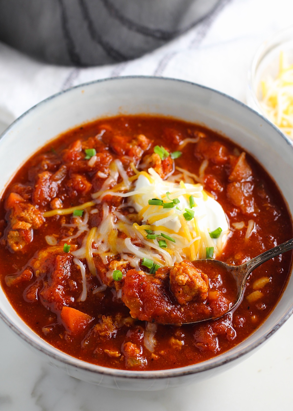 Chicken Chili with Ground Chicken in a bowl with a spoon, topped with sour cream, shredded cheese, and scallions.  On the counter to the right is a bowl with shredded cheese.