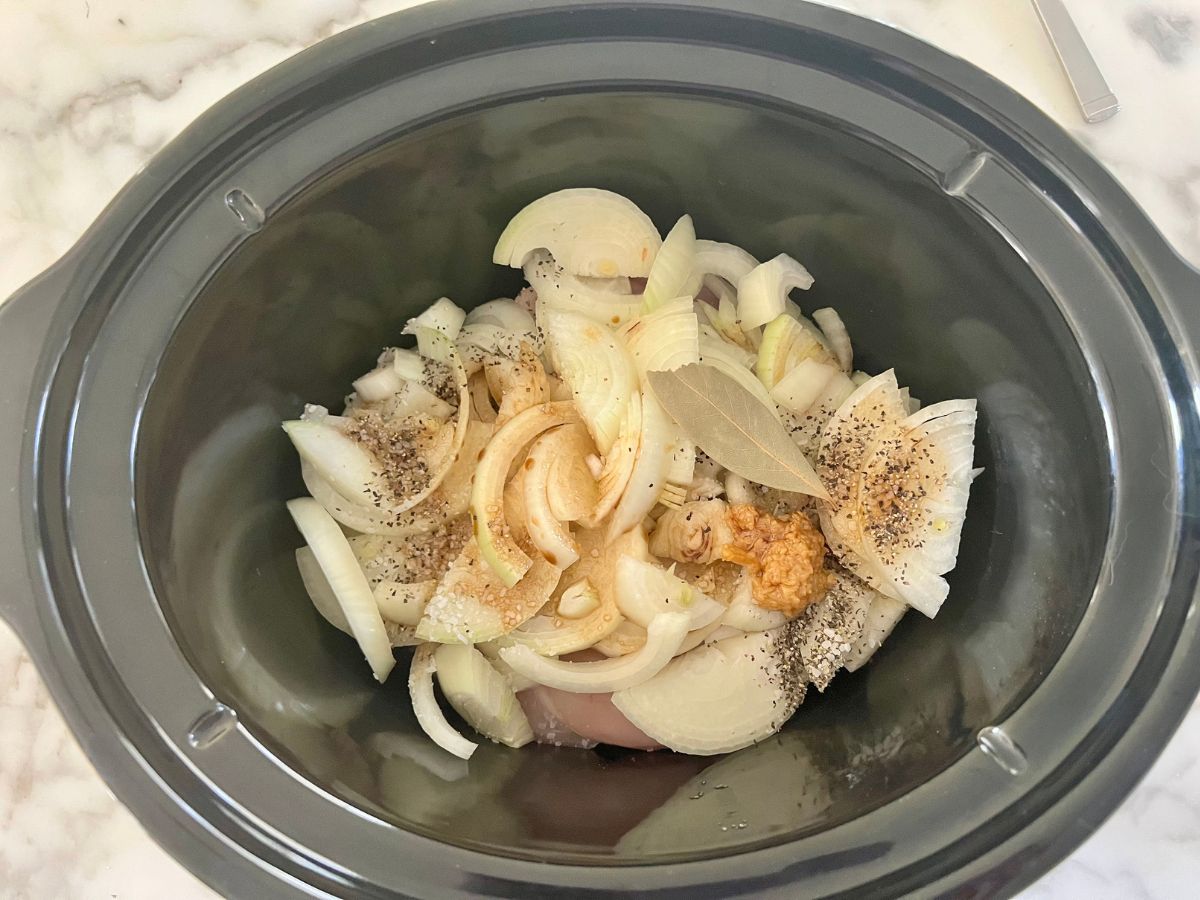 Seasonings, chicken breast and onions added to a crockpot.