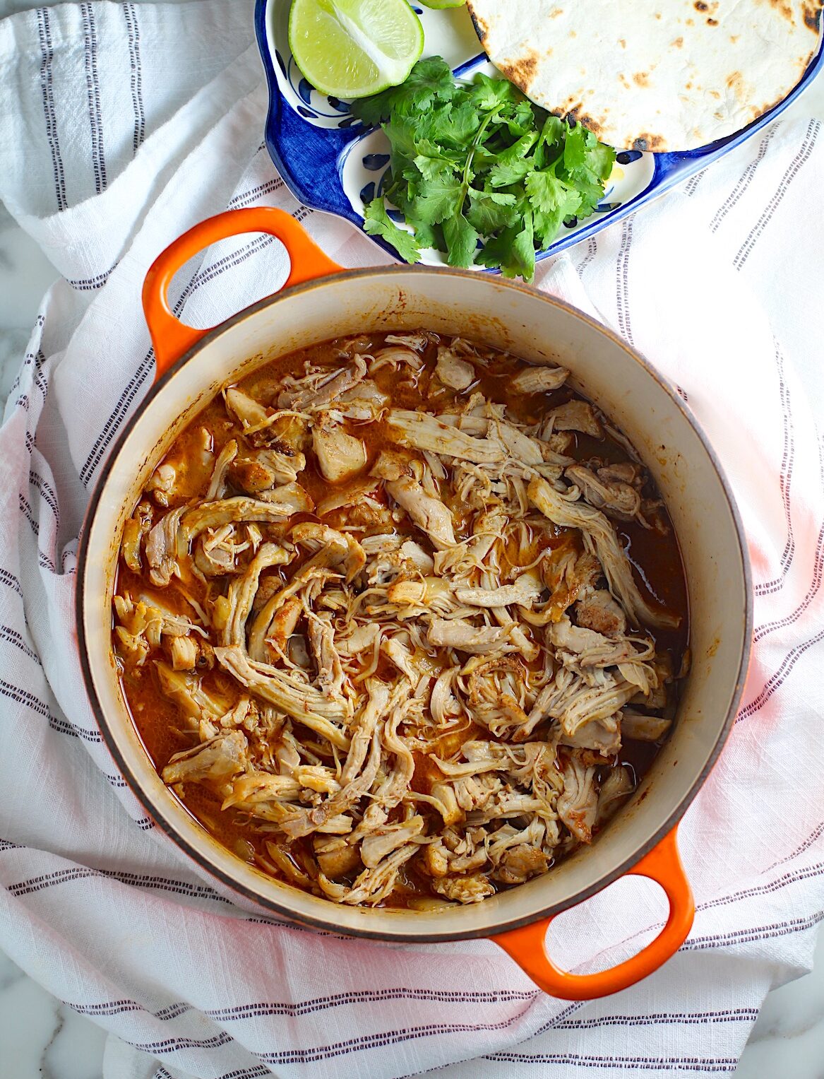 Dutch Oven Pulled Chicken cooked in the dutch oven on a blue and white striped towel with plate of cilantro, lime wedge, and tortillas to the side above in the picture.