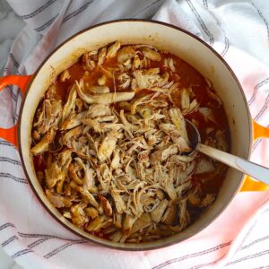 Dutch Oven Pulled Chicken cooked in the dutch oven with a spoon on a blue and white striped towel.