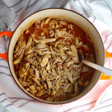 Dutch Oven Pulled Chicken cooked in the dutch oven with a spoon on a blue and white striped towel.