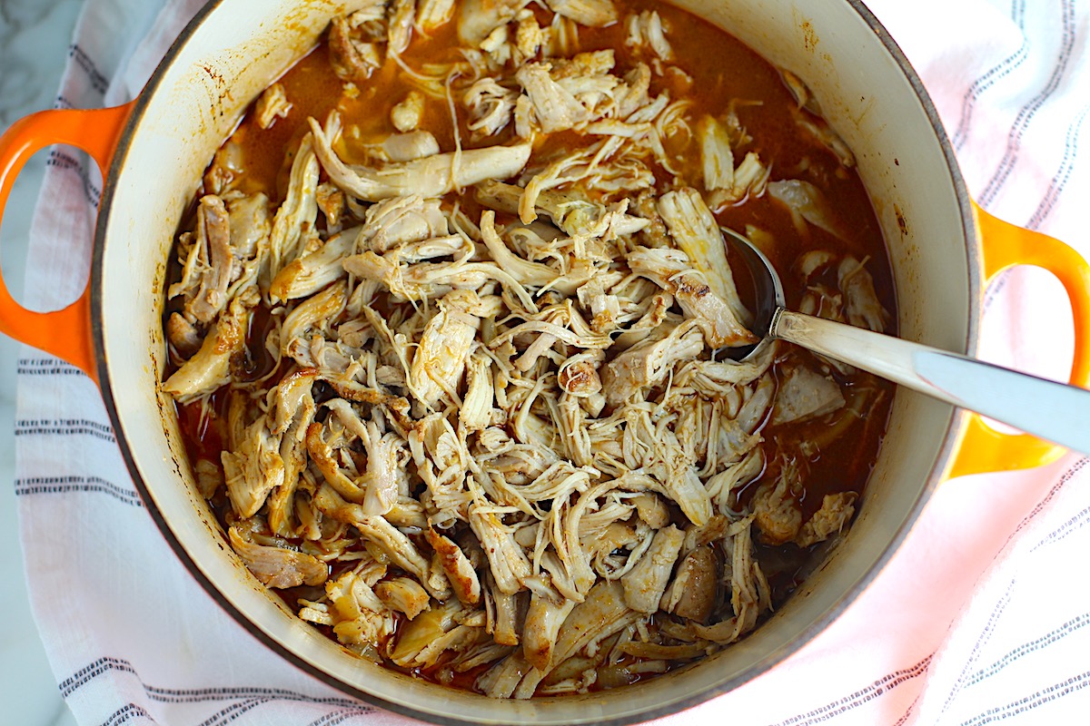 Dutch Oven Pulled Chicken cooked in the dutch oven with a serving spoon on a blue and white striped towel.
