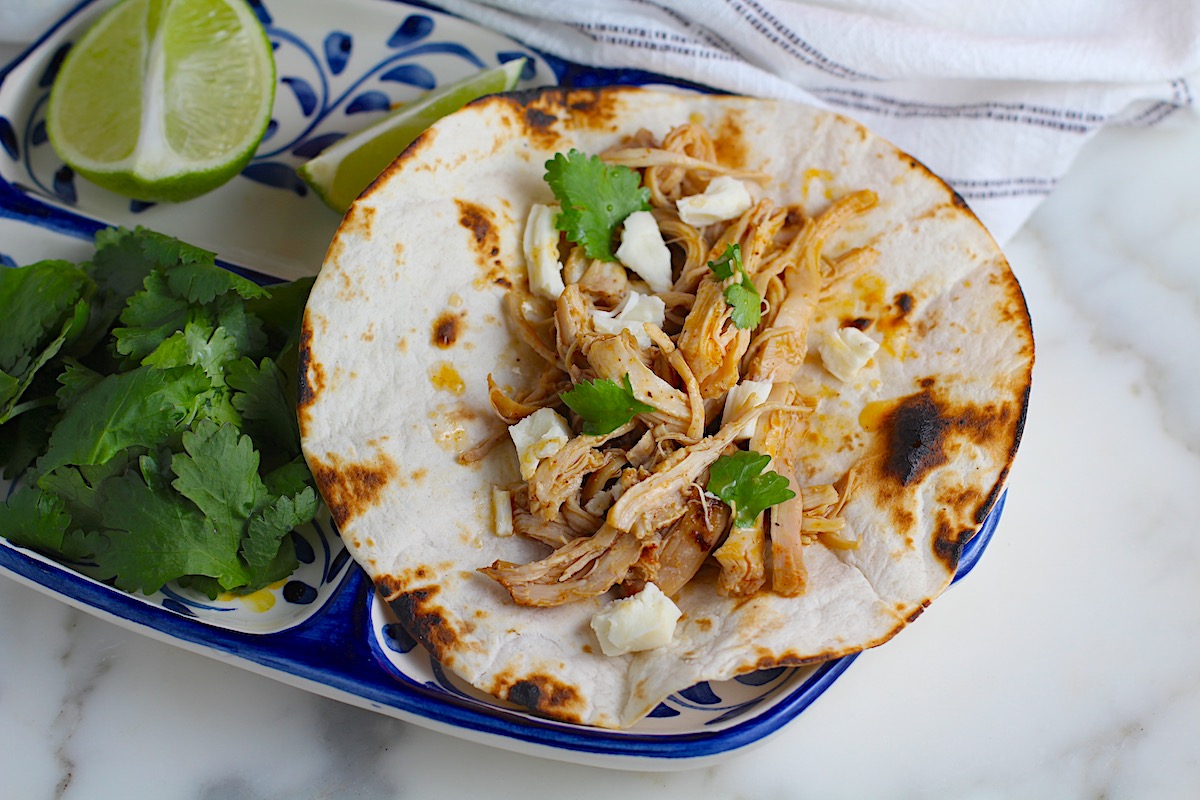 Dutch Oven Pulled Chicken cooked and placed in a grilled flour tortilla with pieces of oaxaca cheese and cilantro on a plate with more cilantro leaves and a lime wedge.