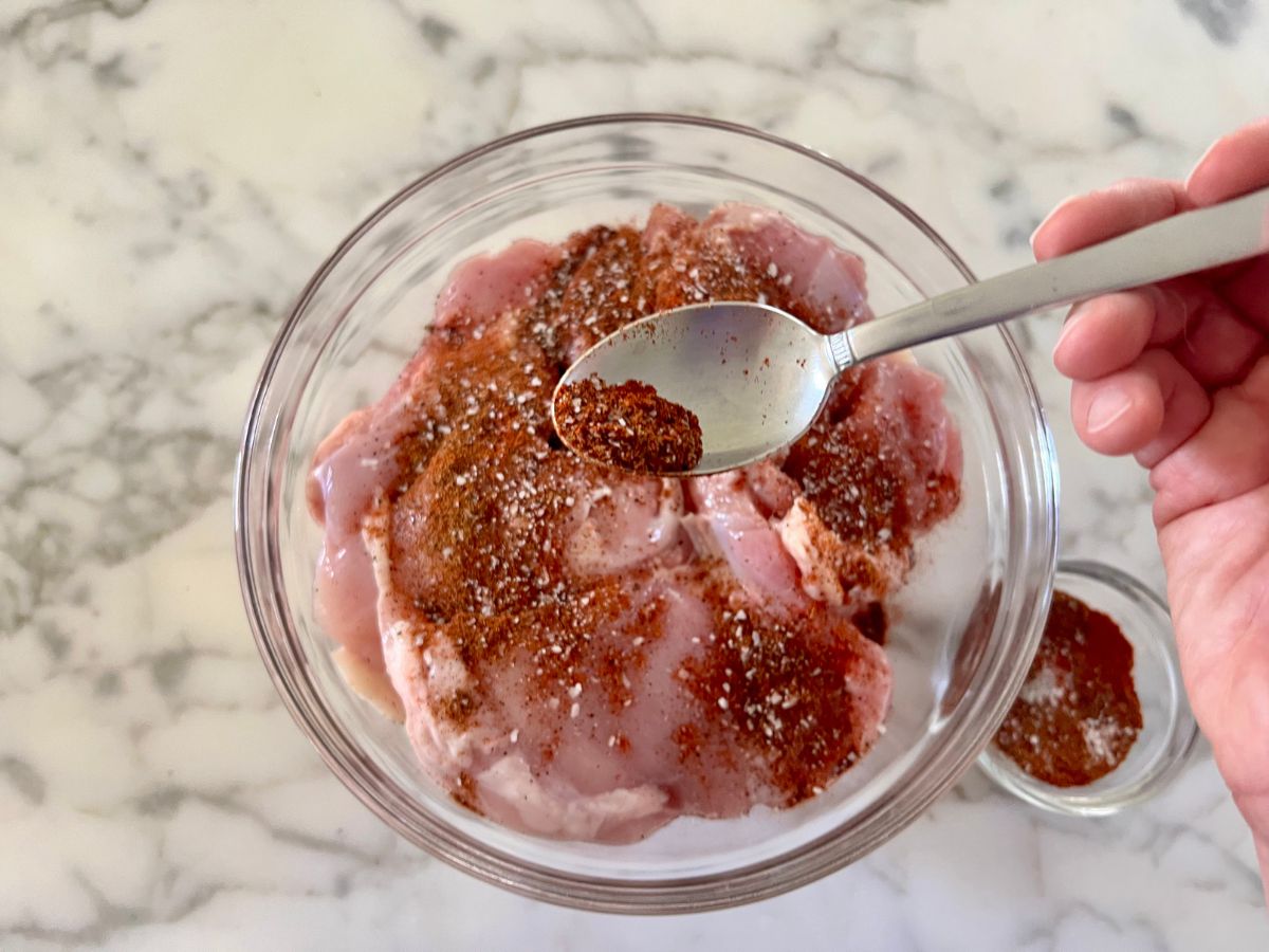 Seasoning mix combined into a small bowl spooned up and sprinkling over a bowl of boneless chicken thighs.