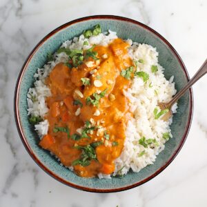 Slow Cooker Satay Chicken with a thick creamy satay sauce over white rice in a bowl garnished with fresh cilantro and chopped peanuts. A spoon is also in the bowl.