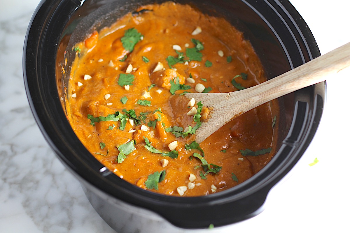 Slow Cooker Satay Chicken in the crockpot insert. It's a thick creamy satay sauce with chicken pieces garnished with fresh cilantro and chopped peanuts with a wood spoon in the pot.