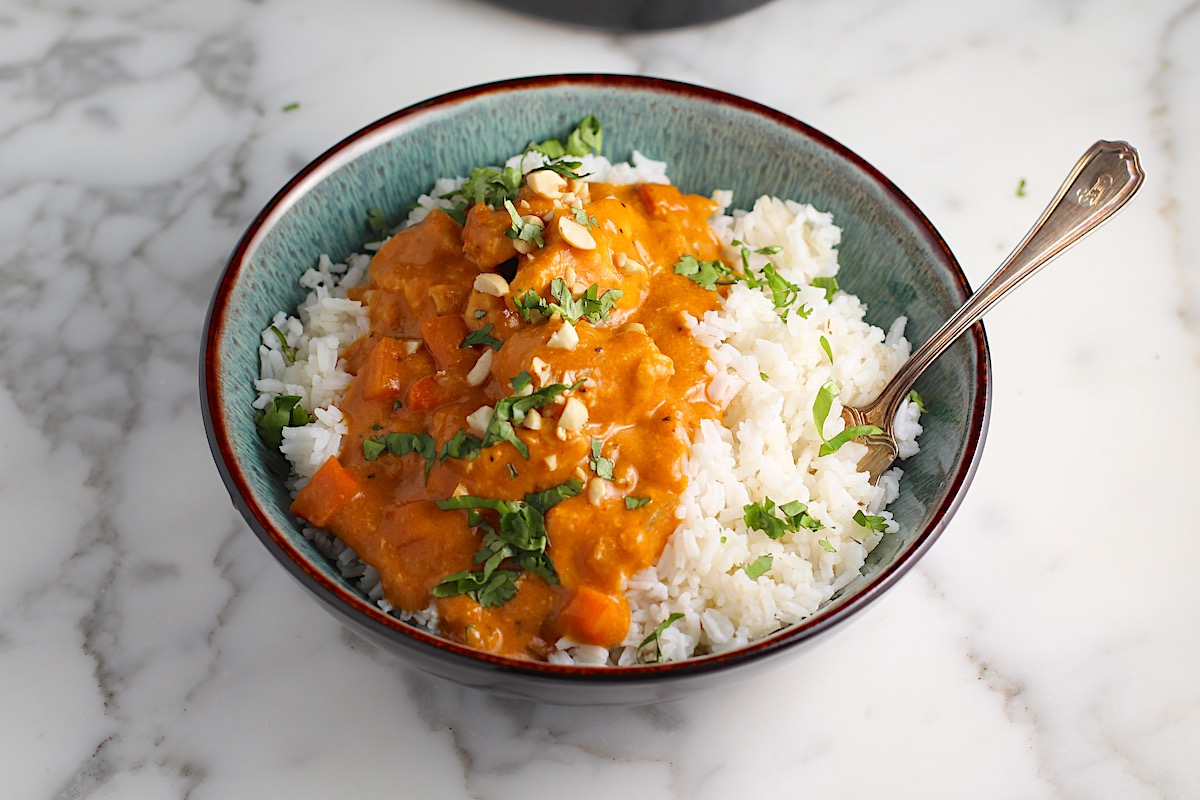 Slow Cooker Satay Chicken with a thick creamy satay sauce over white rice in a bowl garnished with fresh cilantro and chopped peanuts. A spoon is also in the bowl.
