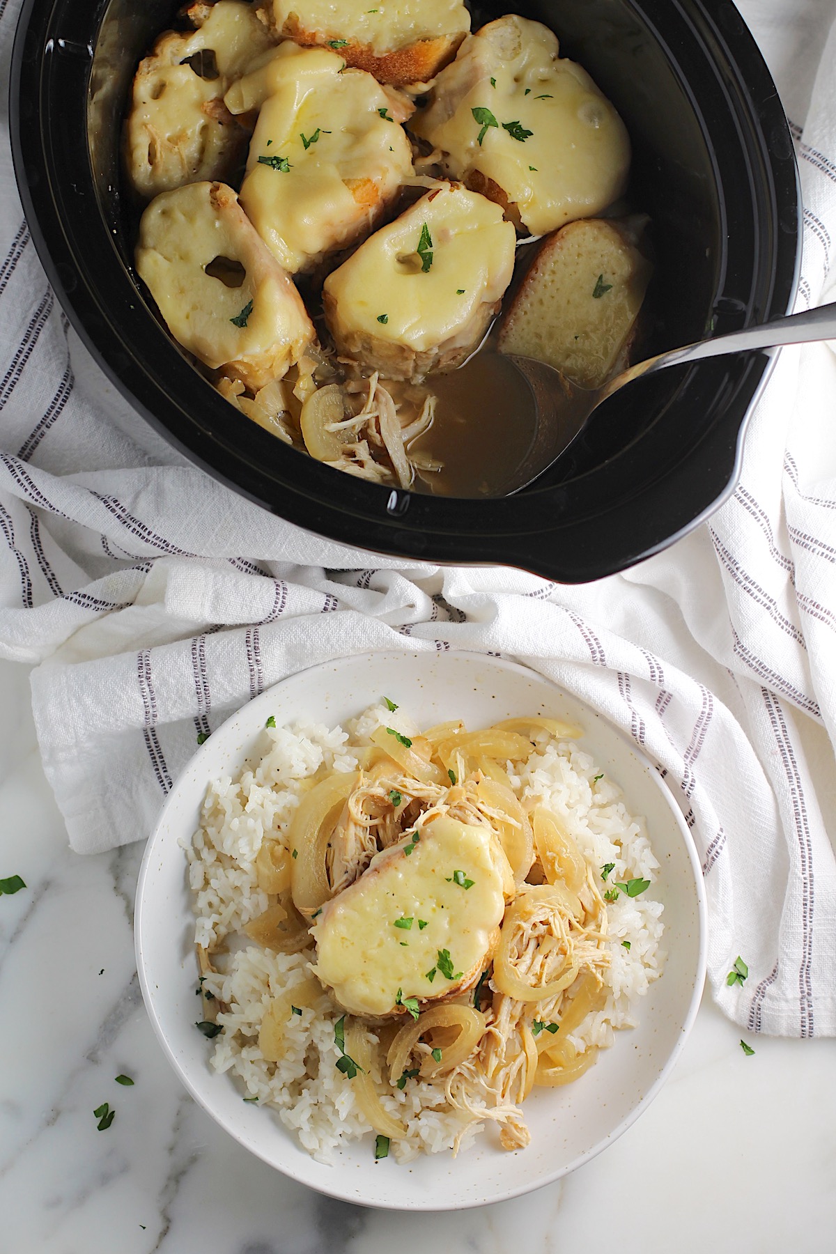 Chicken French Onion Soup Slow Cooker recipe served over cooked white rice on a plate and a slice of baguette on top with melted gruyere cheese chopped fresh parsley. Slow cooker in the background