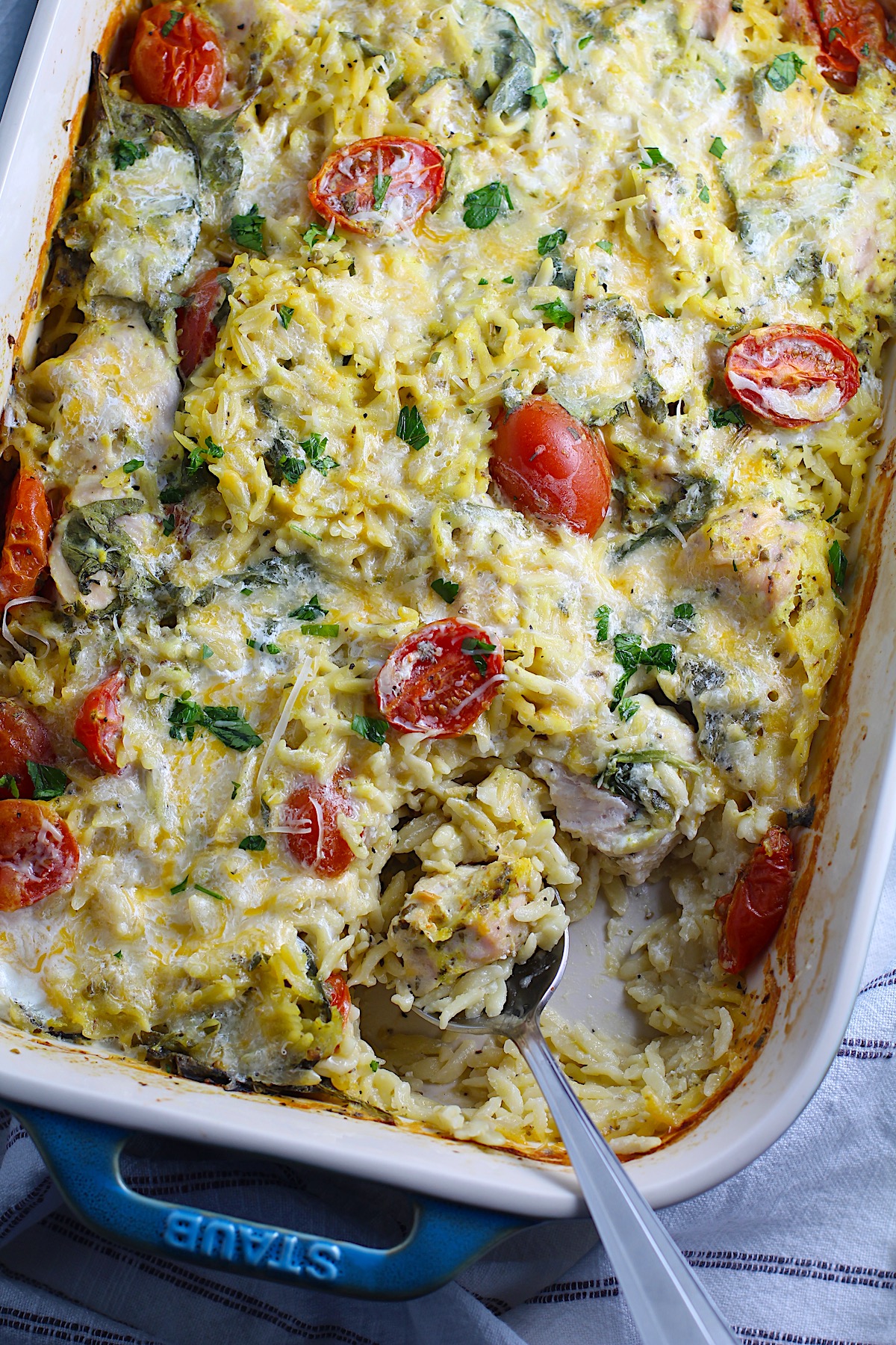 Cheesy Chicken Orzo Bake with grape tomatoes, spinach, and a creamy sauce in a casserole dish with spoon scooping some out at the corner.
