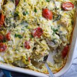 Cheesy Chicken Orzo Bake with grape tomatoes, spinach, and a creamy sauce in a casserole dish with spoon scooping some out at the corner.