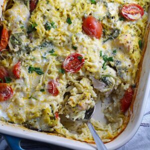 Cheesy Chicken Orzo Bake with grape tomatoes, spinach, and a creamy sauce in a casserole dish with spoon scooping some out at the corner.