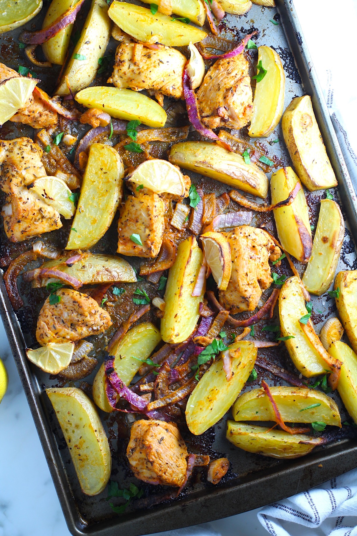 Crispy, golden brown Greek Lemon Chicken and Potatoes with red onions and fresh parsley garnish on a sheet pan on the counter.