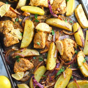 Crispy, golden brown Greek Lemon Chicken and Potatoes with red onions and fresh parsley garnish on a sheet pan on the counter. Whole lemon sitting to the left on the counter.