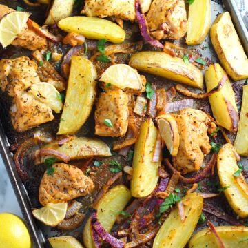 Crispy, golden brown Greek Lemon Chicken and Potatoes with red onions and fresh parsley garnish on a sheet pan on the counter. Whole lemon sitting to the left on the counter.